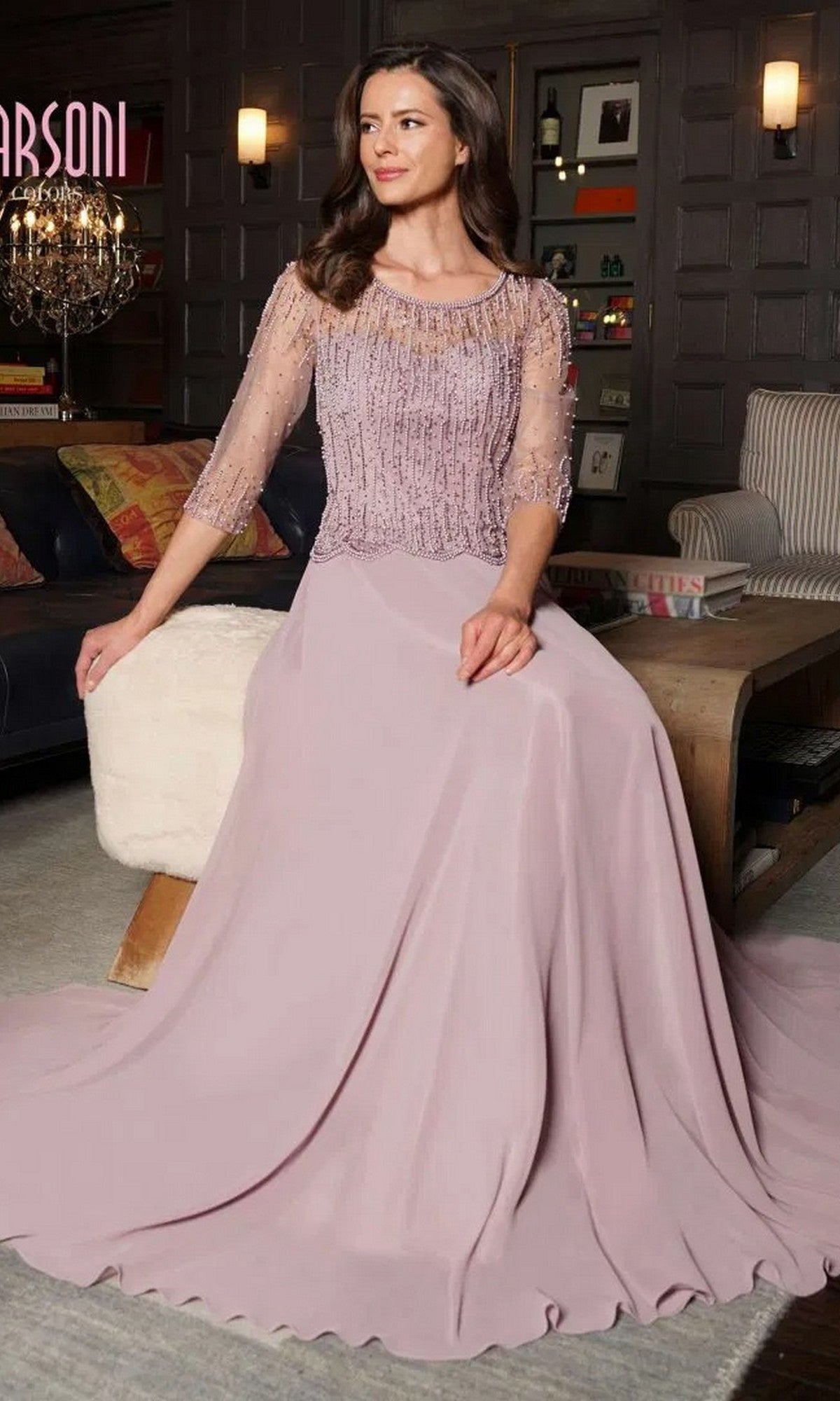 A woman sits on a white stool in a living room, wearing the Marsoni M312 long formal dress in mauve, featuring a beaded bodice and flowing A-line skirt.